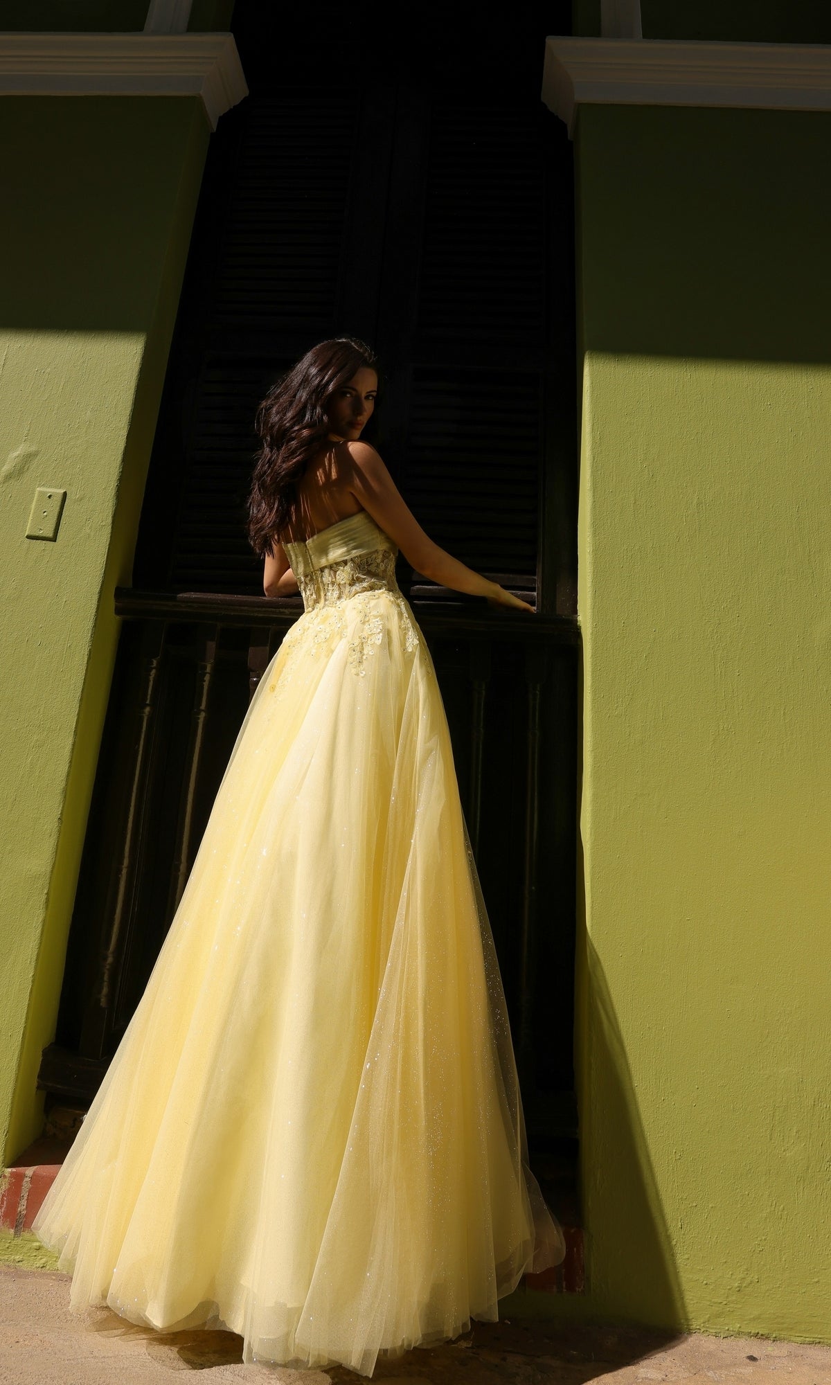 A woman wearing the Strapless Sheer-Waist Long Prom Ball Gown T1326 stands with her back to the camera in front of a green building with dark windows.